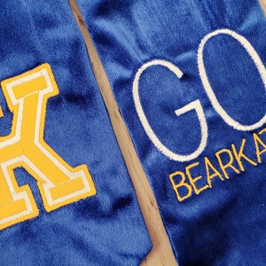 以下が含まれることがあります： 青い卒業式のスツールに、黄色のフェルトの文字「K」と白い刺繍の文字「GO BEARCATS」が施されています。