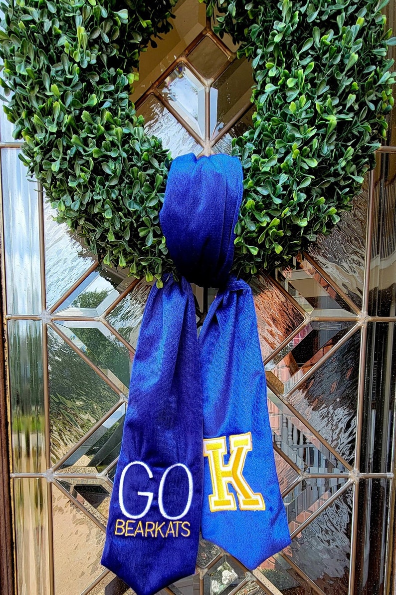 以下が含まれることがあります： 青と金の卒業のタスキに「GO BEARKATS」の文字と反対側に大きな「K」が書かれています。タスキは緑のリースに結ばれています。