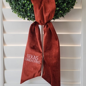 以下が含まれることがあります： 白い糸で「TEXAS LONGHORNS」と刺繍された、マルーンのベルベットのリボン。リボンは緑色のリースに結ばれています。