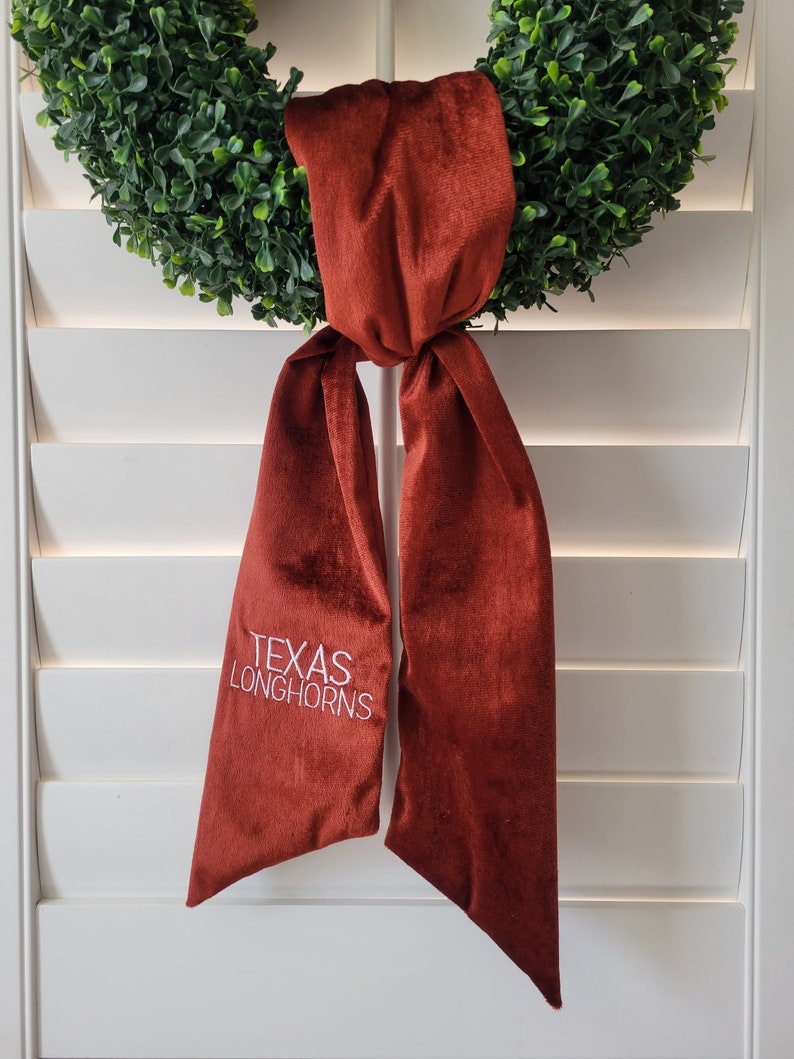 以下が含まれることがあります： 白い糸で「TEXAS LONGHORNS」と刺繍された、マルーンのベルベットのリボン。リボンは緑色のリースに結ばれています。