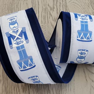 May include: A roll of decorative ribbon with a repeating pattern of blue and white nutcracker soldiers. The ribbon has a white background with the nutcracker design and is bordered by a dark blue velvet trim. Ideal for festive crafts and decorations.