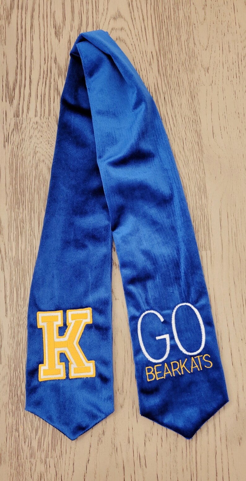 以下が含まれることがあります： 黄色のフェルトの文字「K」と白の刺繍で「GO BEARCATS」と刺繍された青い卒業式ストールです。
