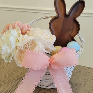 May include: White wicker basket with a pink ribbon that says "Victoria" on it. The basket is filled with pastel coloured Easter eggs and a bouquet of white and pink flowers. A wooden bunny shaped serving tray is behind the basket.