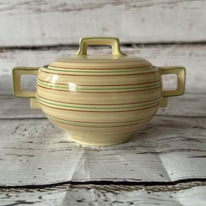 May include: A cream-colored ceramic sugar bowl with a lid. The bowl has green and brown stripes around the body. The lid has a handle on top.