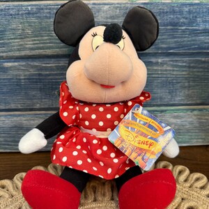 May include: A Minnie Mouse soft toy, wearing a red dress with white polka dots, red shoes, and black ears. The toy has a tag that reads "Applause presents the enchantment of Disney."