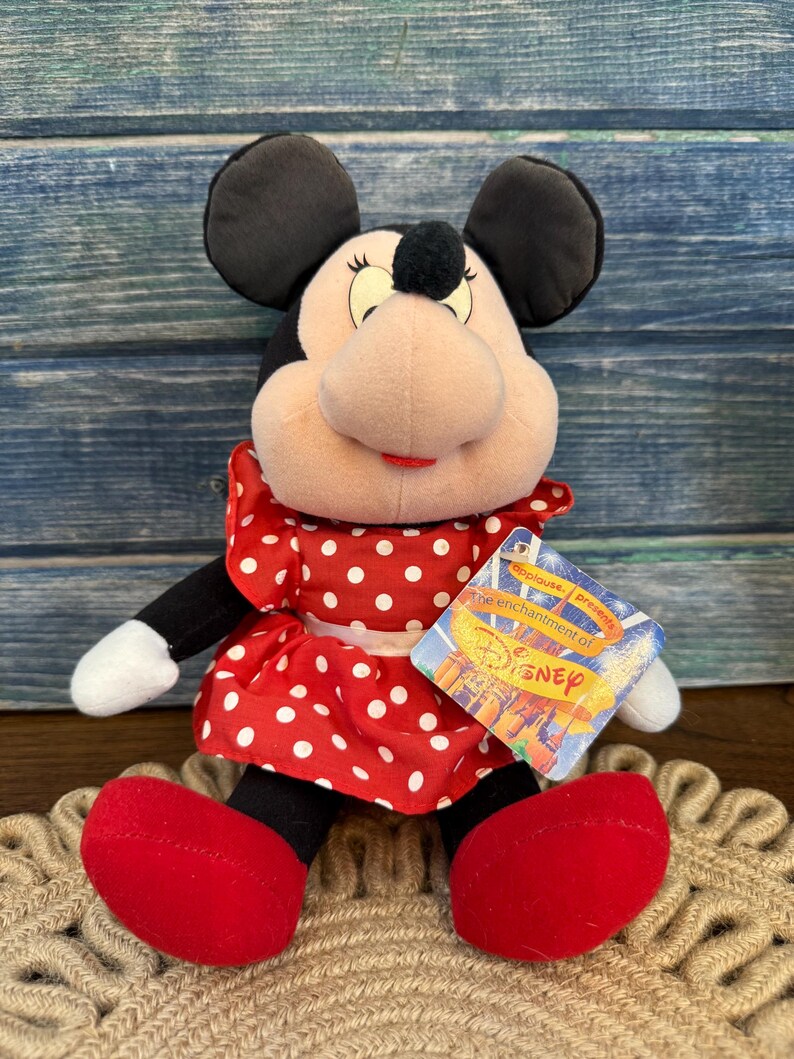 May include: A Minnie Mouse soft toy, wearing a red dress with white polka dots, red shoes, and black ears. The toy has a tag that reads "Applause presents the enchantment of Disney."