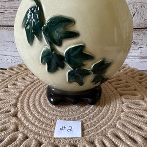 May include: A cream-colored ceramic vase with a green ivy design. The vase has a rounded shape and a black base. The vase is sitting on a brown woven mat with the number "#2" written on a white tag.
