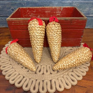 May include: Four decorative pinecone ornaments with gold-colored woven exteriors and red ribbon loops. They are arranged on a round, beige, rope-like placemat. A red wooden box sits in the background.