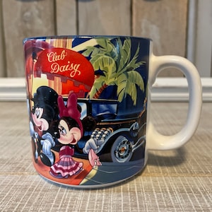 Puede incluir: Una taza de cer&aacute;mica blanca con fondo azul y una imagen colorida de Mickey Mouse y Minnie Mouse frente a un edificio con el texto "Club Daisy".