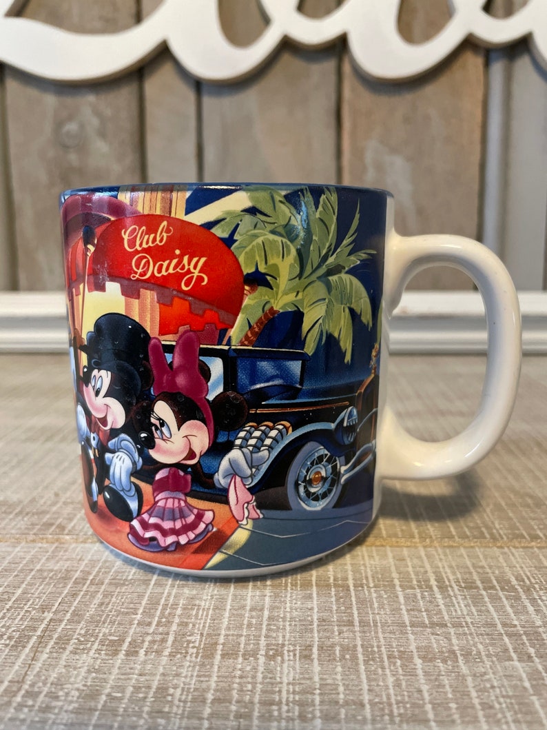 Puede incluir: Una taza de cer&aacute;mica blanca con fondo azul y una imagen colorida de Mickey Mouse y Minnie Mouse frente a un edificio con el texto "Club Daisy".