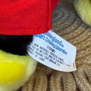 Peut inclure: Gros plan sur une peluche avec un chapeau rouge, des d&eacute;tails noirs et des &eacute;l&eacute;ments jaunes. Une &eacute;tiquette indique "Disneyland Walt Disney World" et "&copy;Disney". L'&eacute;tiquette pr&eacute;cise &eacute;galement les mat&eacute;riaux et l'origine du produit en Chine.
