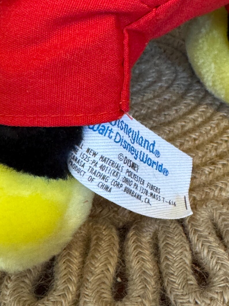 Peut inclure: Gros plan sur une peluche avec un chapeau rouge, des d&eacute;tails noirs et des &eacute;l&eacute;ments jaunes. Une &eacute;tiquette indique "Disneyland Walt Disney World" et "&copy;Disney". L'&eacute;tiquette pr&eacute;cise &eacute;galement les mat&eacute;riaux et l'origine du produit en Chine.