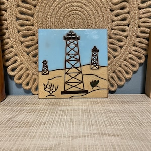 May include: A ceramic tile with a blue background and a brown and white illustration of three oil derricks in a desert landscape.