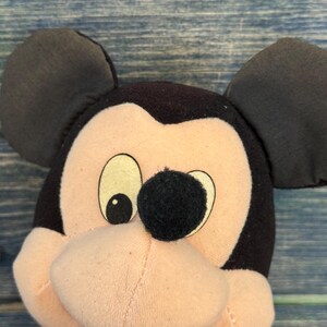 May include: A close-up of a Mickey Mouse soft toy. The toy has black ears, a black nose, and a pink face. It is wearing a blue bow tie and a red vest. The background is a blue wooden surface.