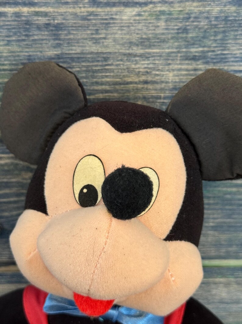 May include: A close-up of a Mickey Mouse soft toy. The toy has black ears, a black nose, and a pink face. It is wearing a blue bow tie and a red vest. The background is a blue wooden surface.