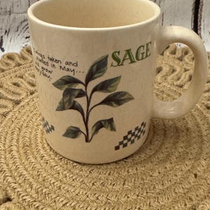 May include: A beige ceramic mug with a sage plant design and the word "Sage" printed on the side. The mug has a black and white checkered pattern around the bottom. The mug is sitting on a woven brown mat.