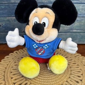 Peut inclure: Une peluche Mickey Mouse avec des oreilles noires, un nez noir et une expression souriante. Mickey porte un t-shirt bleu avec un motif patriotique et un short rouge. Ses pieds jaunes sont visibles, et il est assis sur un tapis tiss&eacute;.