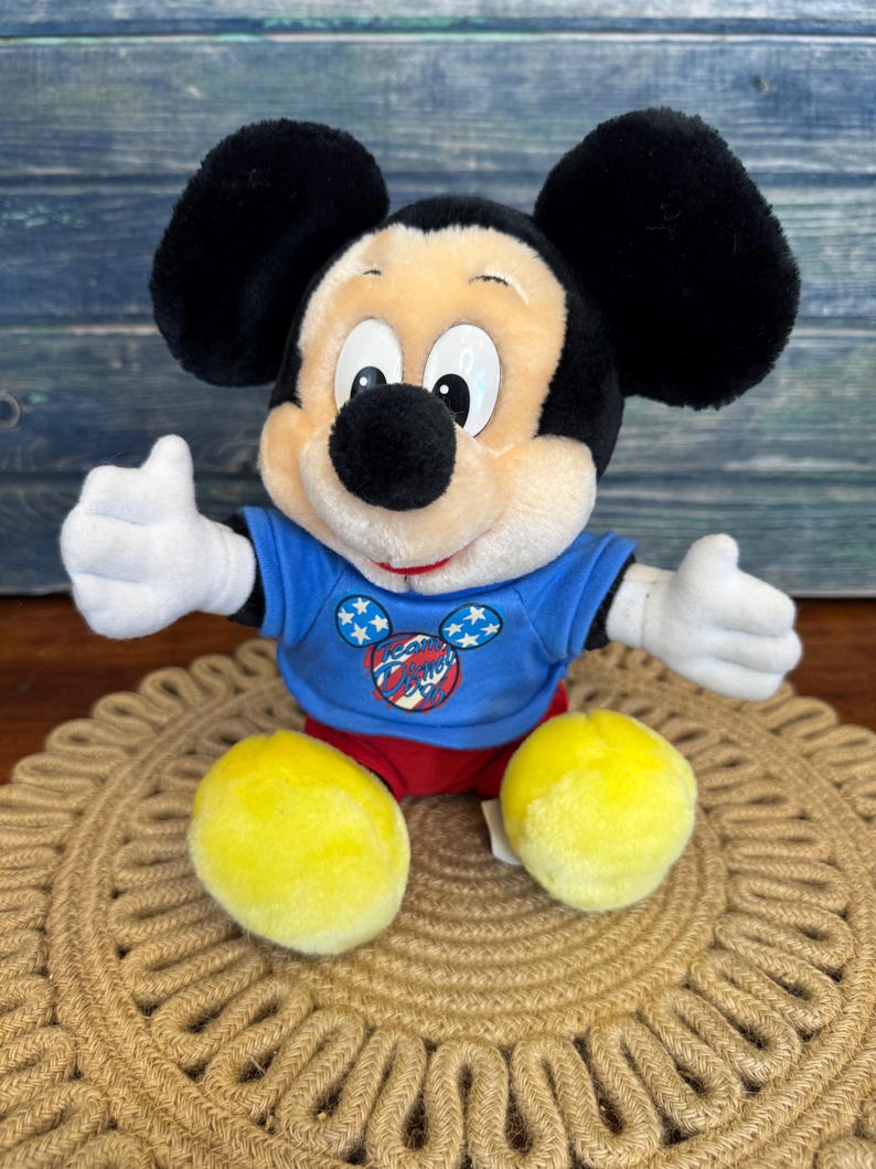 Peut inclure: Une peluche Mickey Mouse avec des oreilles noires, un nez noir et une expression souriante. Mickey porte un t-shirt bleu avec un motif patriotique et un short rouge. Ses pieds jaunes sont visibles, et il est assis sur un tapis tiss&eacute;.