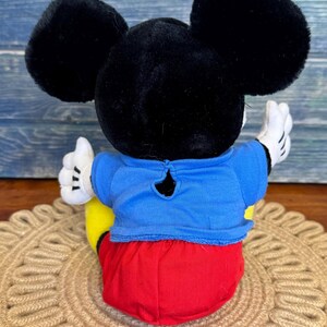 Peut inclure: Une peluche Mickey Mouse, vue de dos, avec de grandes oreilles noires, une chemise bleue, un short rouge et des chaussures jaunes. Le jouet a des gants blancs et est assis sur un tapis rond et tiss&eacute;.