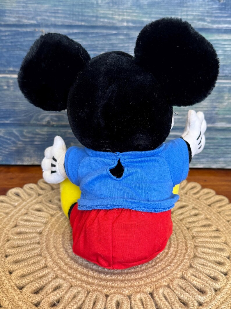Peut inclure: Une peluche Mickey Mouse, vue de dos, avec de grandes oreilles noires, une chemise bleue, un short rouge et des chaussures jaunes. Le jouet a des gants blancs et est assis sur un tapis rond et tiss&eacute;.