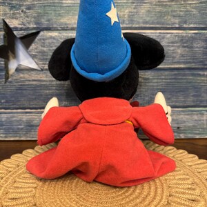 Puede incluir: Un peluche de un rat&oacute;n negro con un sombrero de mago azul con estrellas blancas y una t&uacute;nica roja. El juguete est&aacute; sentado sobre una estera tejida de color beige. El fondo es una superficie de madera azul.