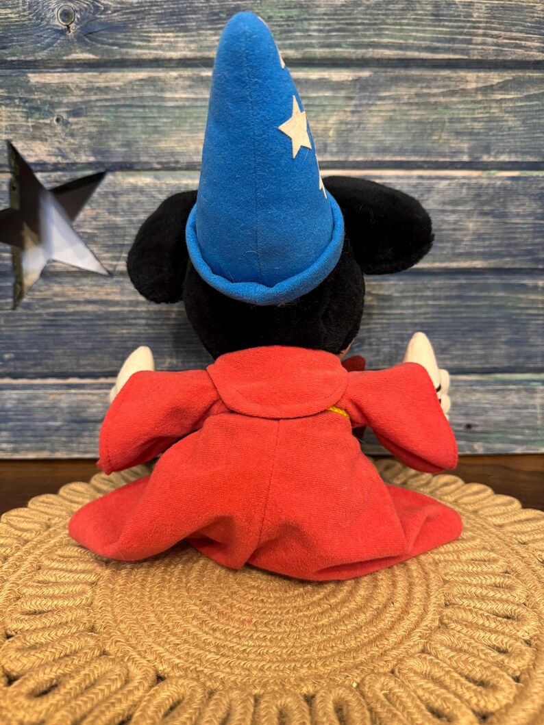 Puede incluir: Un peluche de un rat&oacute;n negro con un sombrero de mago azul con estrellas blancas y una t&uacute;nica roja. El juguete est&aacute; sentado sobre una estera tejida de color beige. El fondo es una superficie de madera azul.