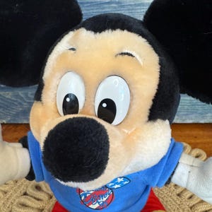 Peut inclure: Une poup&eacute;e en peluche de Mickey Mouse avec de grandes oreilles noires, un visage bronz&eacute; et de grands yeux blancs. Mickey porte une chemise bleue avec un logo Disney, un short rouge et des chaussures jaunes. La poup&eacute;e est assise sur une surface tiss&eacute;e.
