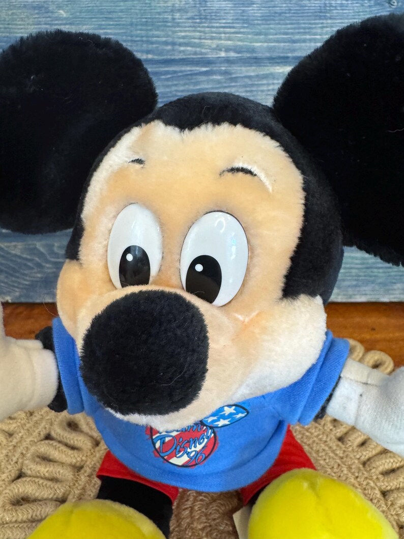 Peut inclure: Une poup&eacute;e en peluche de Mickey Mouse avec de grandes oreilles noires, un visage bronz&eacute; et de grands yeux blancs. Mickey porte une chemise bleue avec un logo Disney, un short rouge et des chaussures jaunes. La poup&eacute;e est assise sur une surface tiss&eacute;e.
