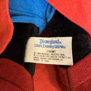 Puede incluir: Primer plano de un peluche con una chaqueta roja, una camisa azul y una cara negra. Una etiqueta blanca dice "Disneyland Walt Disney World" y "&copy; Disney". La etiqueta tambi&eacute;n enumera los materiales y la informaci&oacute;n de fabricaci&oacute;n.