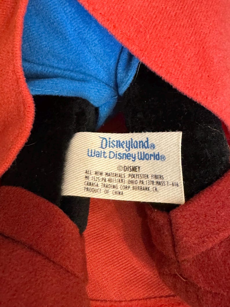 Puede incluir: Primer plano de un peluche con una chaqueta roja, una camisa azul y una cara negra. Una etiqueta blanca dice "Disneyland Walt Disney World" y "&copy; Disney". La etiqueta tambi&eacute;n enumera los materiales y la informaci&oacute;n de fabricaci&oacute;n.