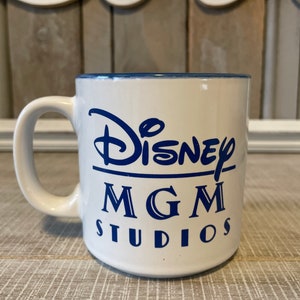 Puede incluir: Taza de cer&aacute;mica blanca con borde azul y letras azules que dicen "Disney MGM Studios".