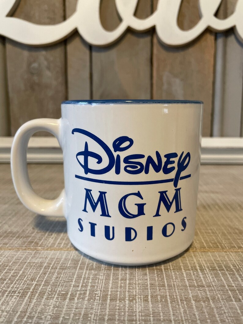 Puede incluir: Taza de cer&aacute;mica blanca con borde azul y letras azules que dicen "Disney MGM Studios".