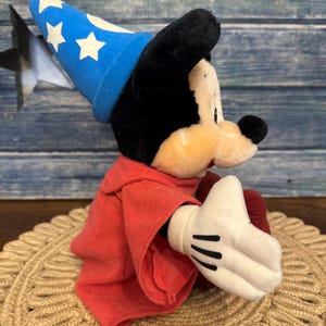 Puede incluir: Un peluche de Mickey Mouse vestido de hechicero, con un sombrero azul con estrellas blancas y una t&uacute;nica roja. El juguete tiene orejas negras, una mano enguantada blanca y est&aacute; sentado sobre una estera tejida.