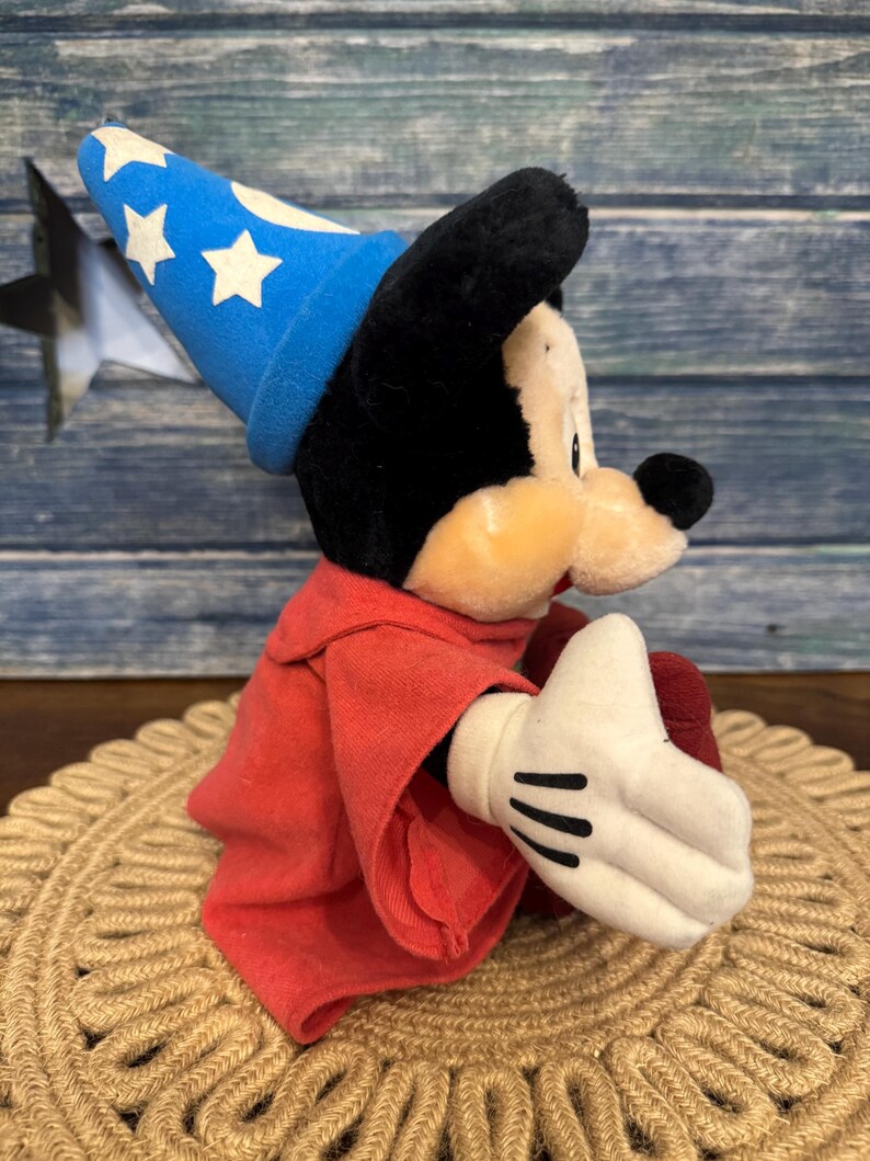 Puede incluir: Un peluche de Mickey Mouse vestido de hechicero, con un sombrero azul con estrellas blancas y una t&uacute;nica roja. El juguete tiene orejas negras, una mano enguantada blanca y est&aacute; sentado sobre una estera tejida.