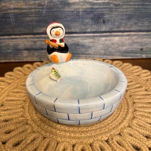 May include: A ceramic decorative bowl with a light blue igloo design. A small penguin figurine wearing a red hat and scarf sits on the bowl's edge, holding a fishing rod. A small fish figurine is inside the bowl. The bowl rests on a brown woven mat.