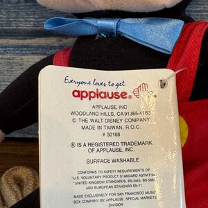 May include: A soft toy of a cartoon character, featuring a black body, red shorts, and a blue bow tie. The attached tag reads "Everyone loves to get applause." The tag also includes the company's address and trademark information.