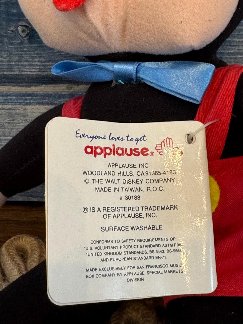 May include: A soft toy of a cartoon character, featuring a black body, red shorts, and a blue bow tie. The attached tag reads "Everyone loves to get applause." The tag also includes the company's address and trademark information.