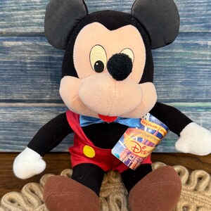 May include: A plush Mickey Mouse doll with black ears, a black nose, and a red bow tie. The doll is wearing red shorts with yellow buttons and brown shoes. A Disney tag is attached to the doll.