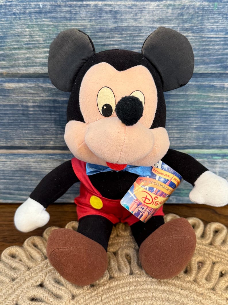 May include: A Mickey Mouse soft toy with black ears, a black nose, and a red bow tie. The toy is wearing red shorts with yellow buttons and brown shoes. A Disney tag is attached to the toy.