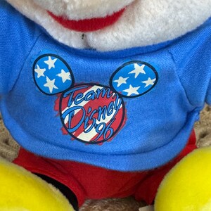 Peut inclure: Une peluche portant un t-shirt bleu avec le texte "Team Disney '96" et des motifs d'oreilles de Mickey Mouse avec des &eacute;toiles blanches. Le jouet a un short rouge, des pieds jaunes et un visage blanc avec un sourire rouge.
