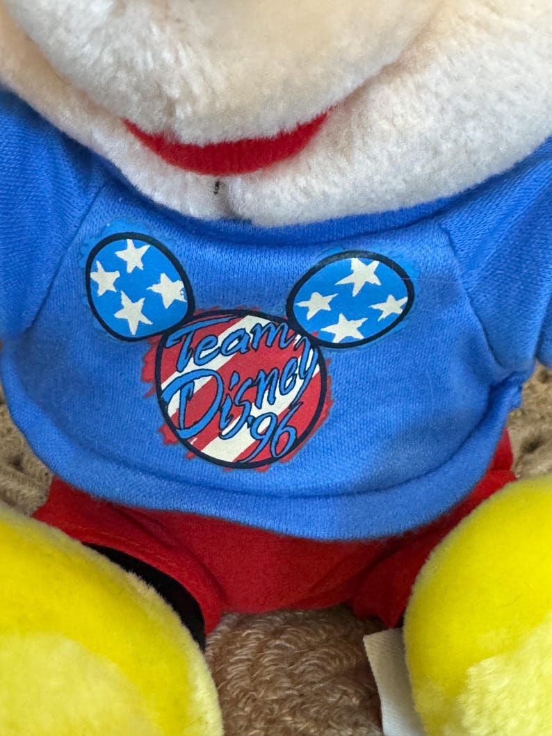 Peut inclure: Une peluche portant un t-shirt bleu avec le texte "Team Disney '96" et des motifs d'oreilles de Mickey Mouse avec des &eacute;toiles blanches. Le jouet a un short rouge, des pieds jaunes et un visage blanc avec un sourire rouge.