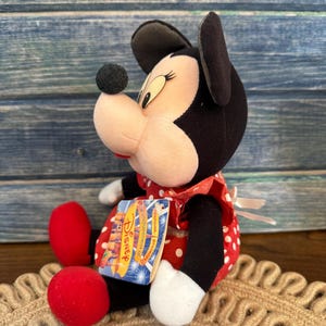 May include: A plush Minnie Mouse doll, with a black head and ears, a pink face, and a red and white polka dot dress. The doll has red shoes and a Disney tag. The doll is sitting on a woven mat.