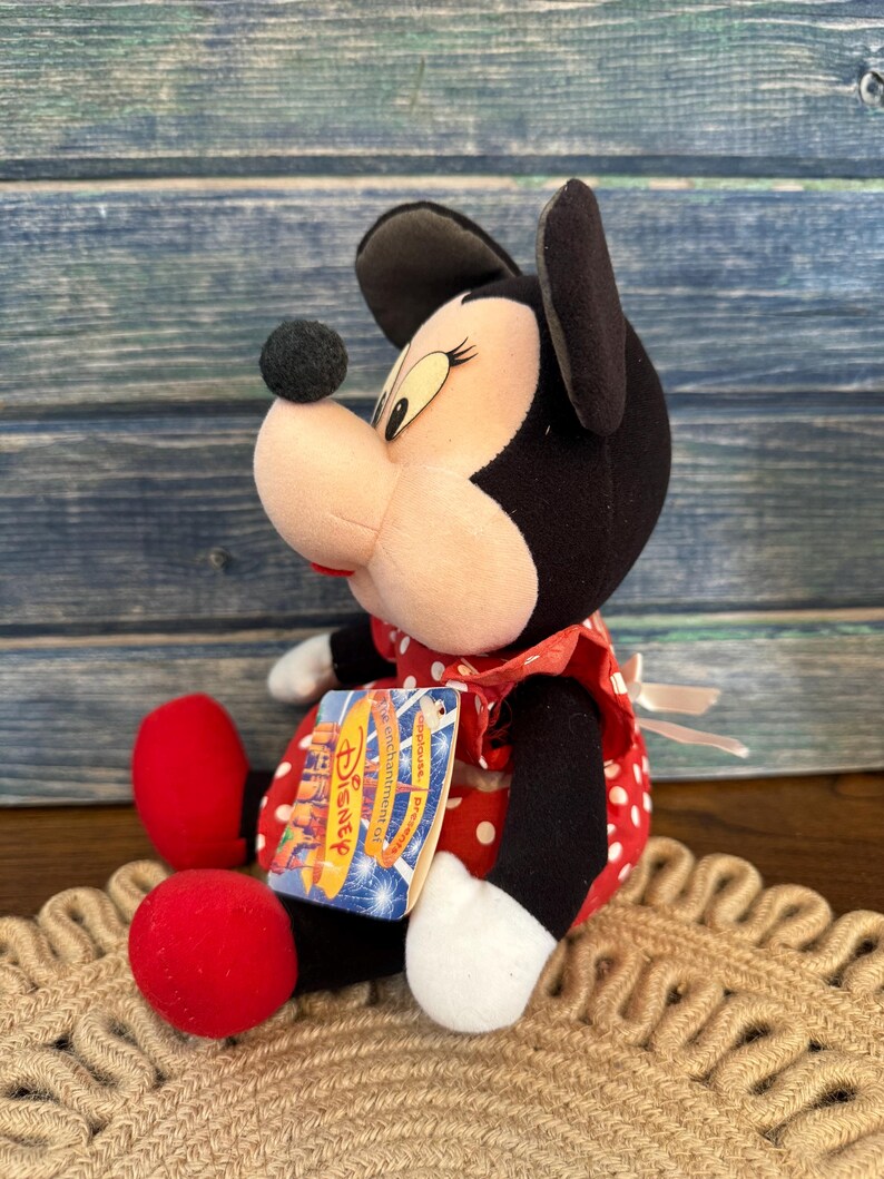 May include: A plush Minnie Mouse doll, with a black head and ears, a pink face, and a red and white polka dot dress. The doll has red shoes and a Disney tag. The doll is sitting on a woven mat.