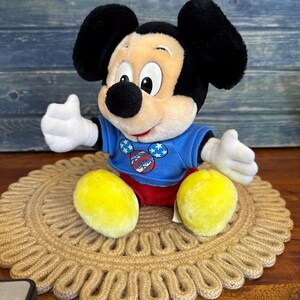 Peut inclure: Une peluche Mickey Mouse portant un t-shirt bleu avec un motif patriotique, un short rouge et des chaussures jaunes. La poup&eacute;e est assise sur un dessous de plat rond et tiss&eacute;. L'arri&egrave;re-plan est une surface en bois bleu.