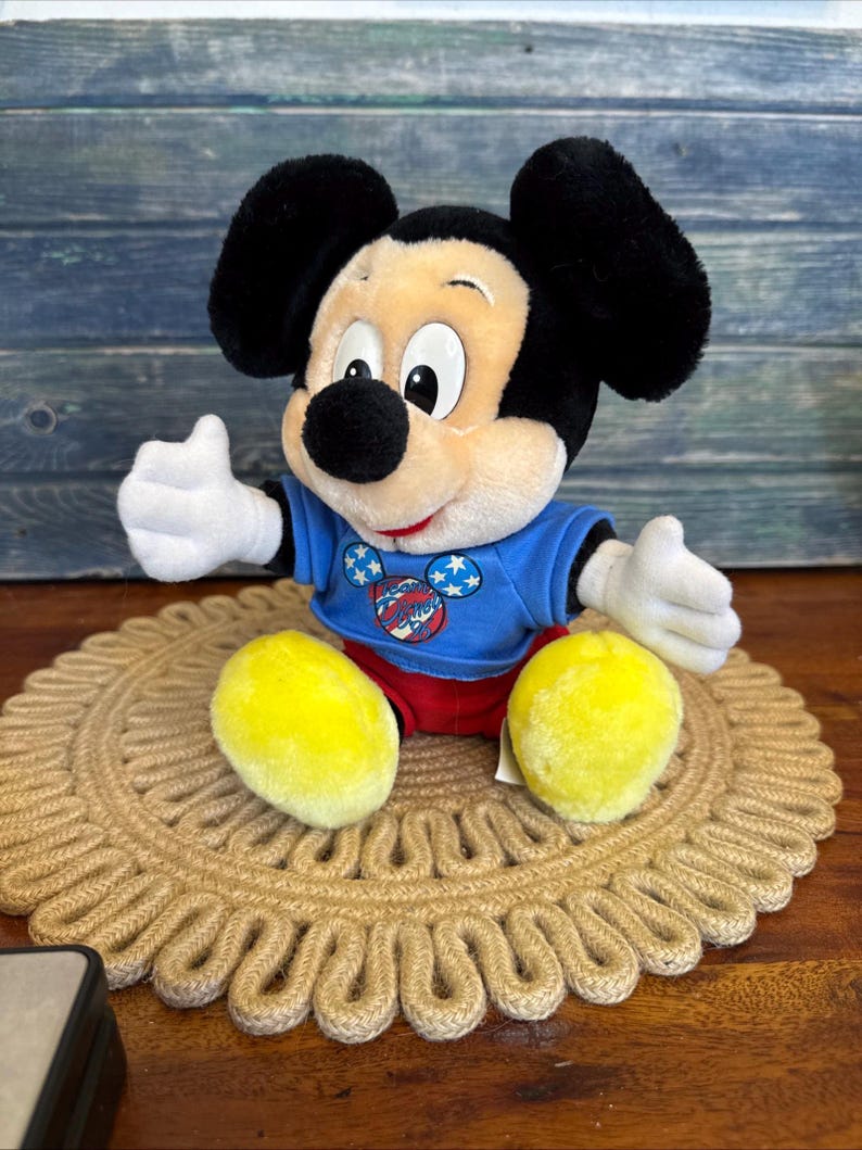 Peut inclure: Une peluche Mickey Mouse portant un t-shirt bleu avec un motif patriotique, un short rouge et des chaussures jaunes. La poup&eacute;e est assise sur un dessous de plat rond et tiss&eacute;. L'arri&egrave;re-plan est une surface en bois bleu.