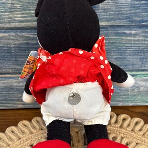 May include: A Minnie Mouse soft toy, seen from behind, with black ears and head, a red dress with white spots, white bloomers, and red shoes. The doll has a tag attached and is on a woven mat.