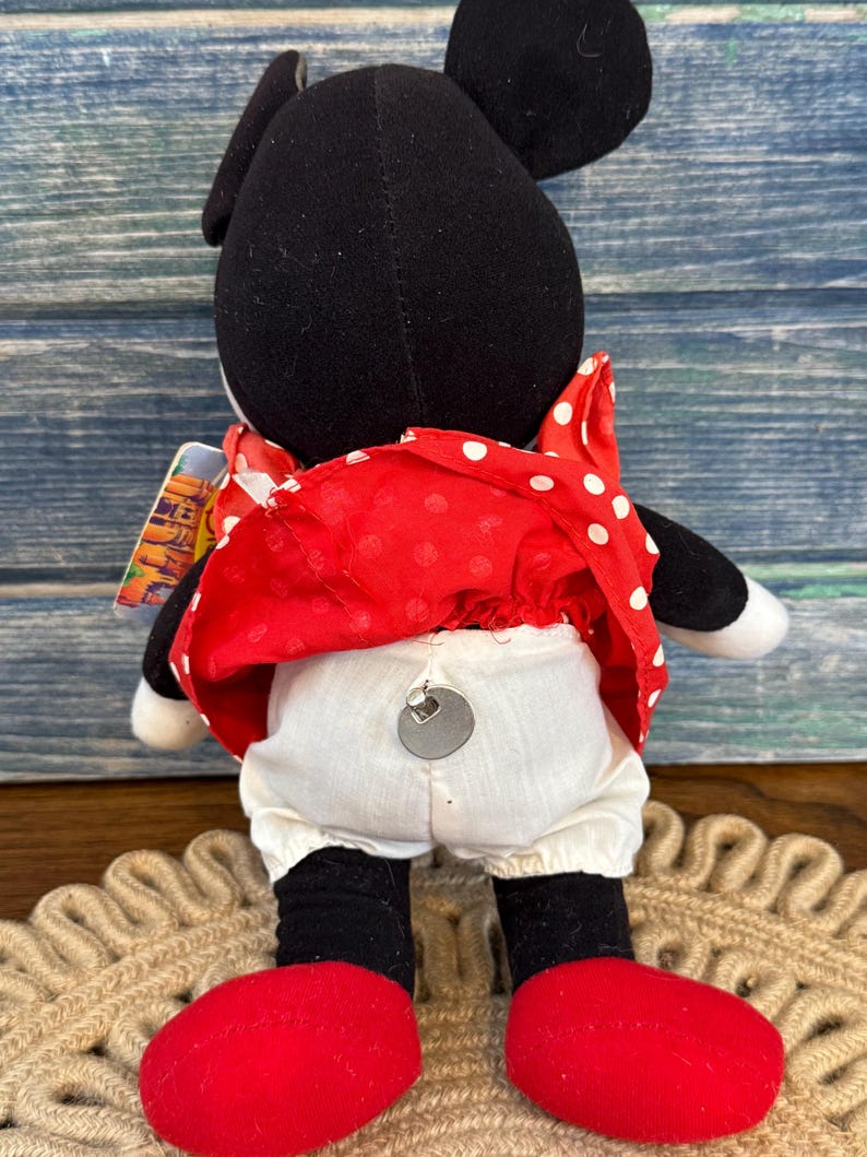 May include: A Minnie Mouse soft toy, seen from behind, with black ears and head, a red dress with white spots, white bloomers, and red shoes. The doll has a tag attached and is on a woven mat.