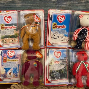 May include: Collection of Ty Beanie Baby bears in their original packaging. The bears are dressed in various country-themed outfits, including a bear with a German flag, a bear with an American flag, and a red bear with a Mexican flag. The packaging includes the text "International Bears II".