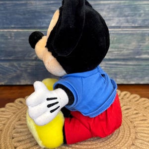 Peut inclure: Une peluche Mickey Mouse, avec des oreilles et une t&ecirc;te noires, un visage beige et une chemise bleue. Le jouet porte un pantalon rouge, des chaussures jaunes et des gants blancs &agrave; rayures noires. Le jouet est assis sur un tapis tress&eacute;.