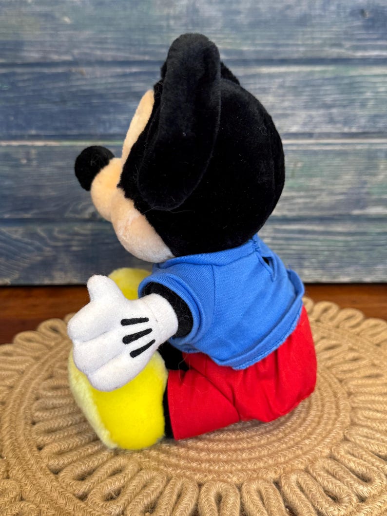 Peut inclure: Une peluche Mickey Mouse, avec des oreilles et une t&ecirc;te noires, un visage beige et une chemise bleue. Le jouet porte un pantalon rouge, des chaussures jaunes et des gants blancs &agrave; rayures noires. Le jouet est assis sur un tapis tress&eacute;.